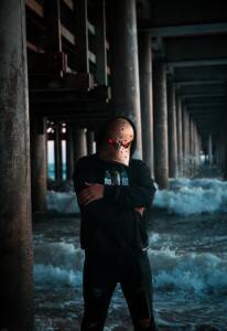 Image: Dark background under a bridge with a bad guy wearing a scary mask with arms folded.