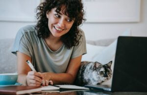 glad woman with cat writing in planner while using laptop